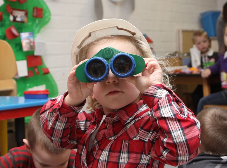 Kind kijkt op de kinderopvang door verrekijker naar de camera.