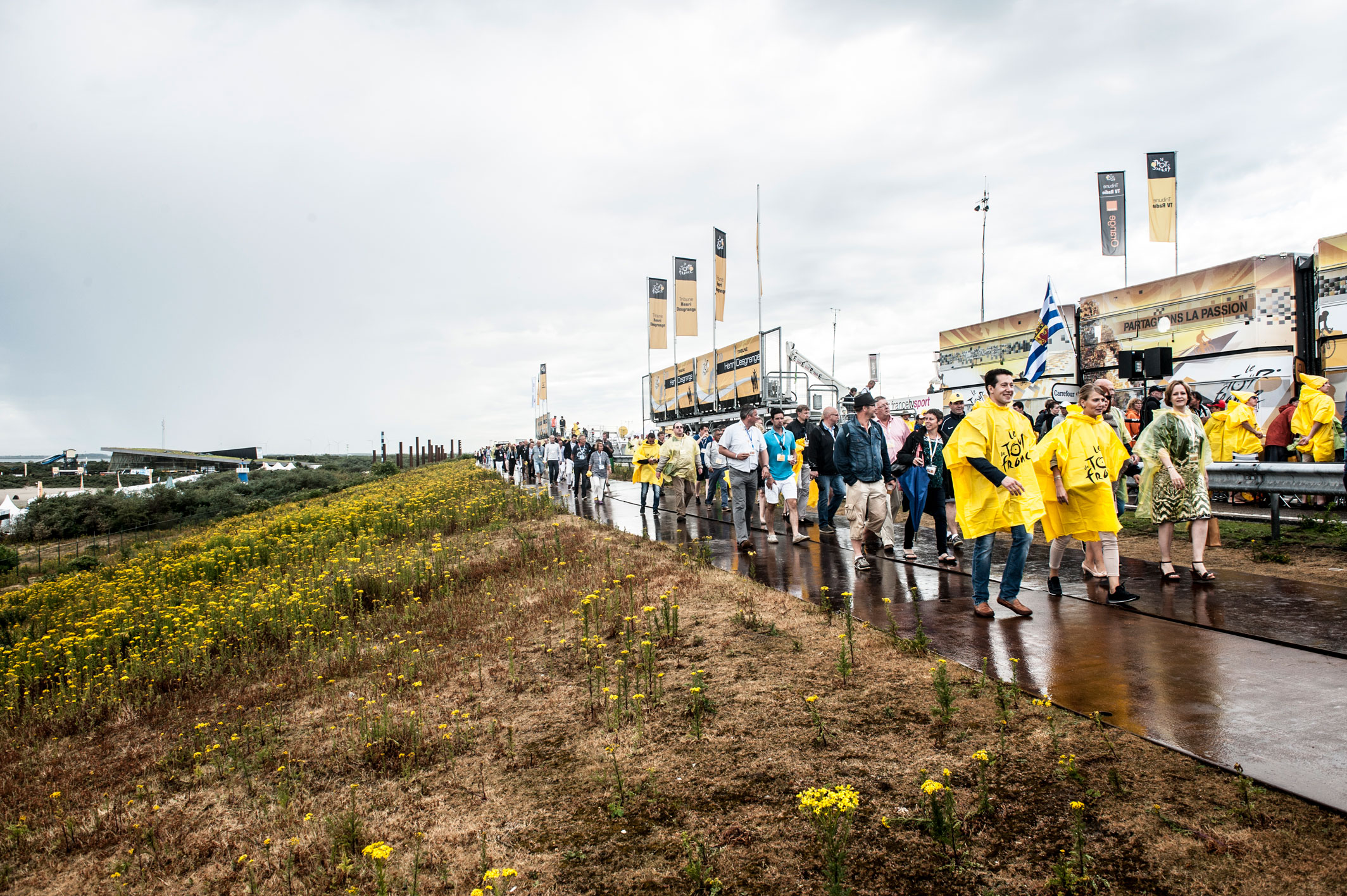 Toeschouwers van de Tour de France lopen op de Oosterscheldekering in een gele poncho in de regen